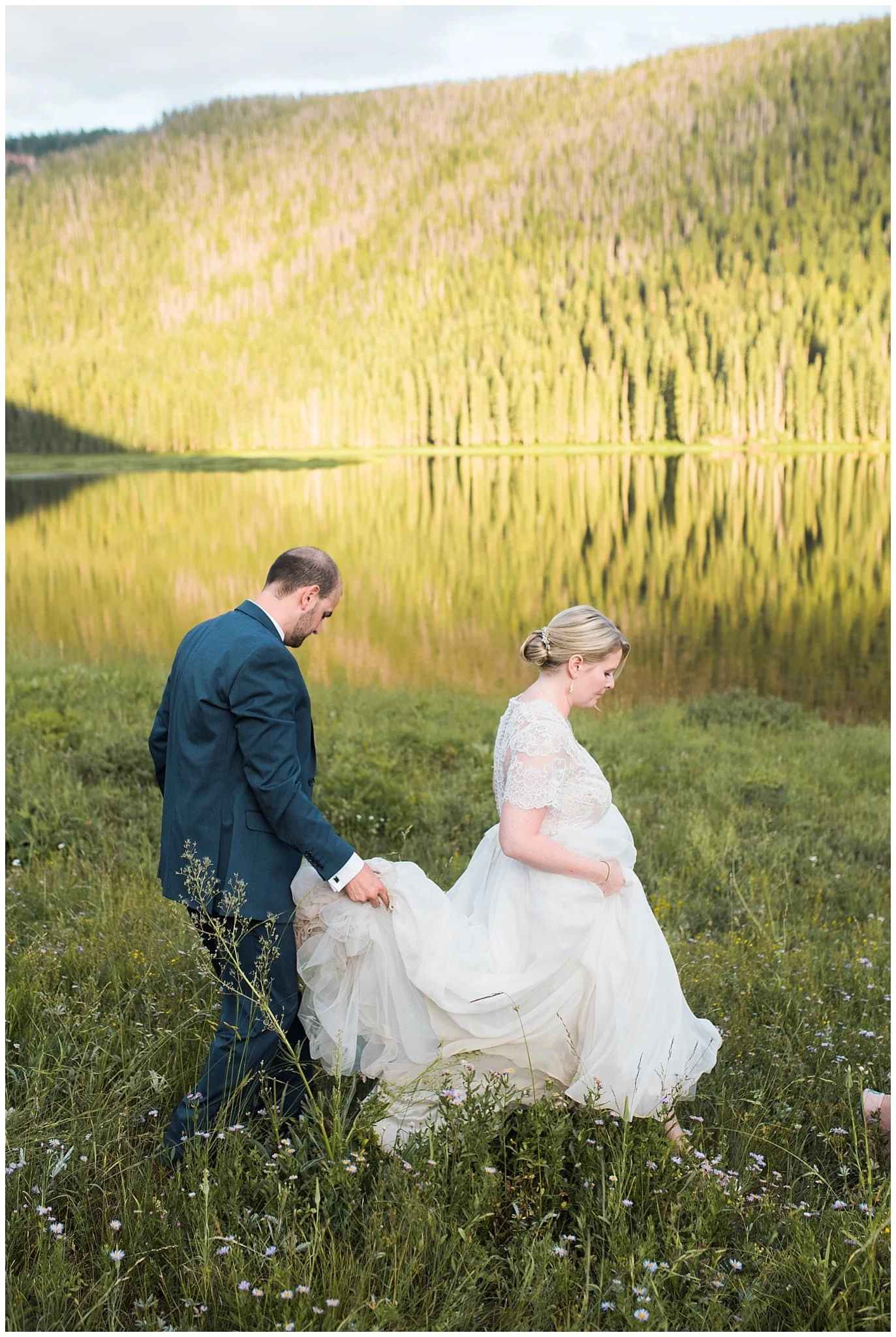 bride in lace dress and groom holding train walking by lake at Piney River Ranch wedding by Aspen wedding photographer Jennie Crate photographer