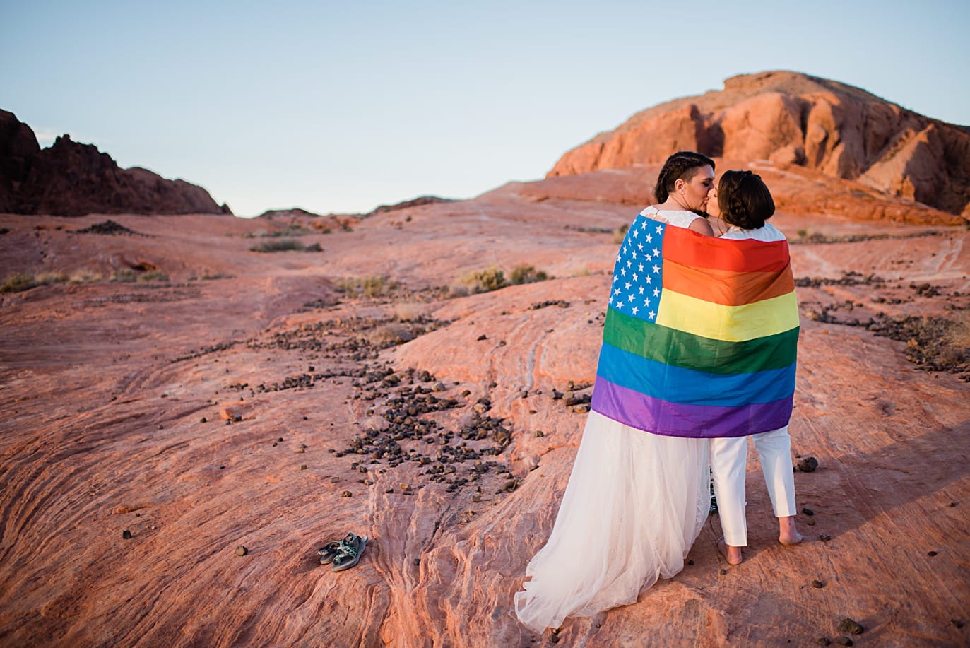 two brides wrapped in pride flag on wedding day at sunset in Valley of Fire Nevada at Valley of Fire wedding by Las Vegas wedding photographer Jennie Crate