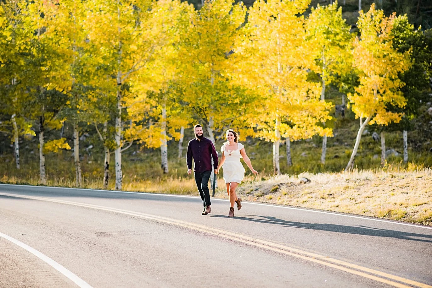 couple runs across the road in front of golden aspens at RMNP engagement session by Boudler engagement photographer Jennie Crate