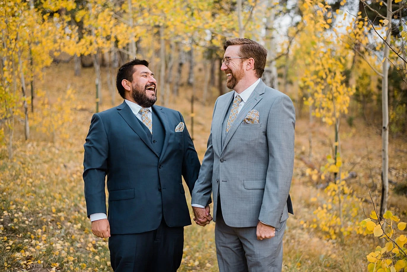 two grooms in navy and gray suits laugh in fall aspen trees in Golden Gate Canyon by Colorado gay wedding photographer Jennie Crate