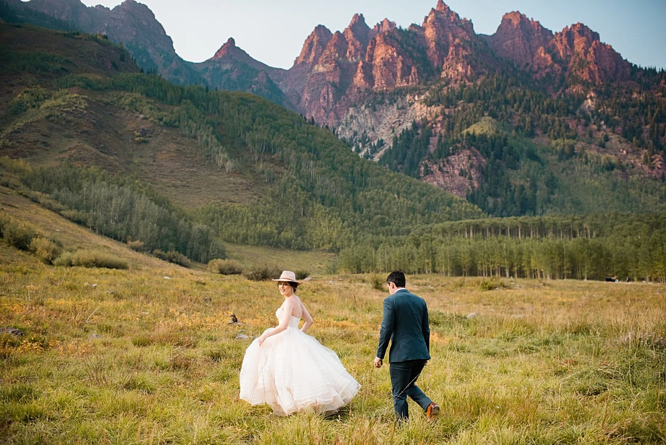 bride in pink hat walks in front of red rocks at fall Maroon Bells wedding by Snowmass wedding photographer Jennie Crate