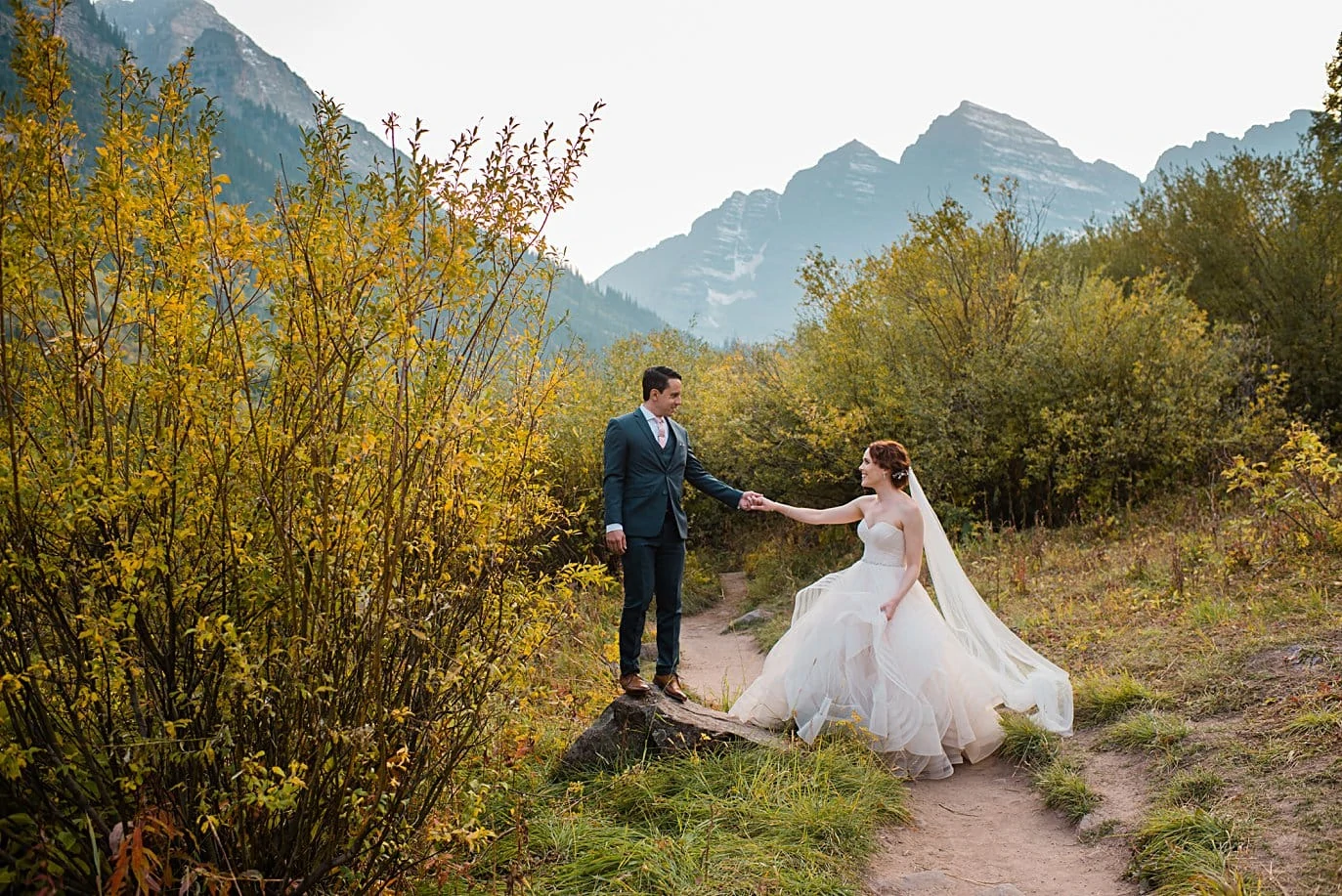 groom helps bride up on rock at fall Maroon Bells wedding by Snowmass wedding photographer Jennie Crate
