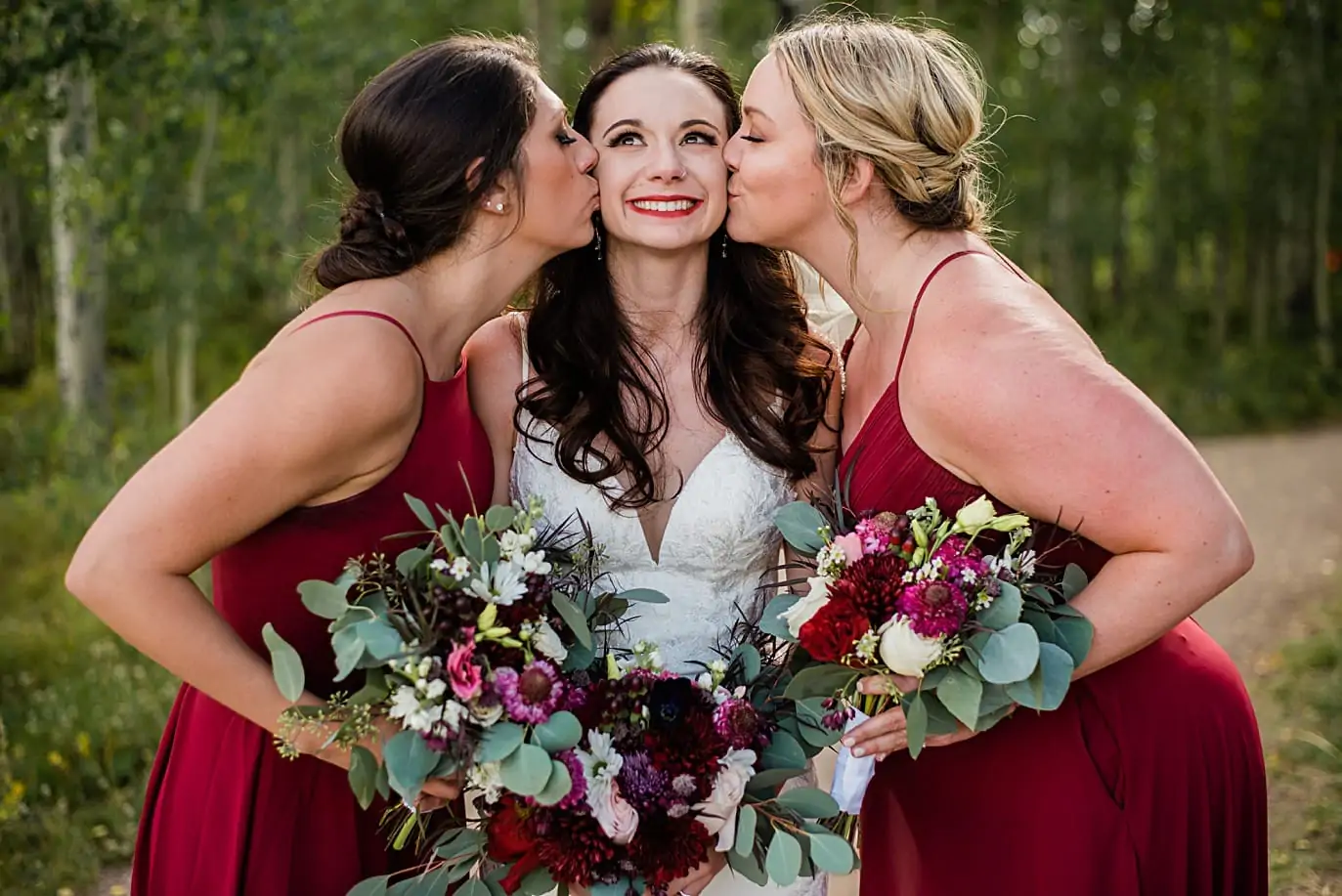 two bridesmaids kiss bride on cheek prior to her wedding ceremony on the top of a mountain