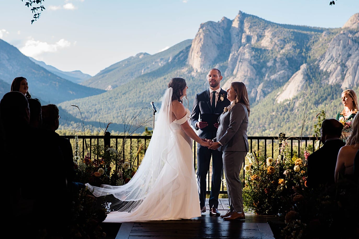two brides stand in front of balcony and view of Estes Park rocky peaks during ceremony at The Boulders wedding