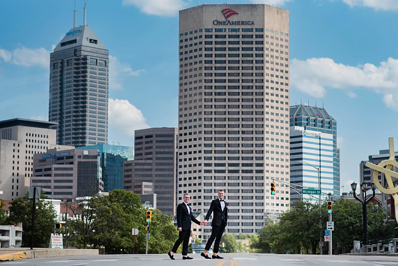 two grooms in tuxedos walk hand in hand across the downtown indianaplois skyline before their gay wedding in indianapolis